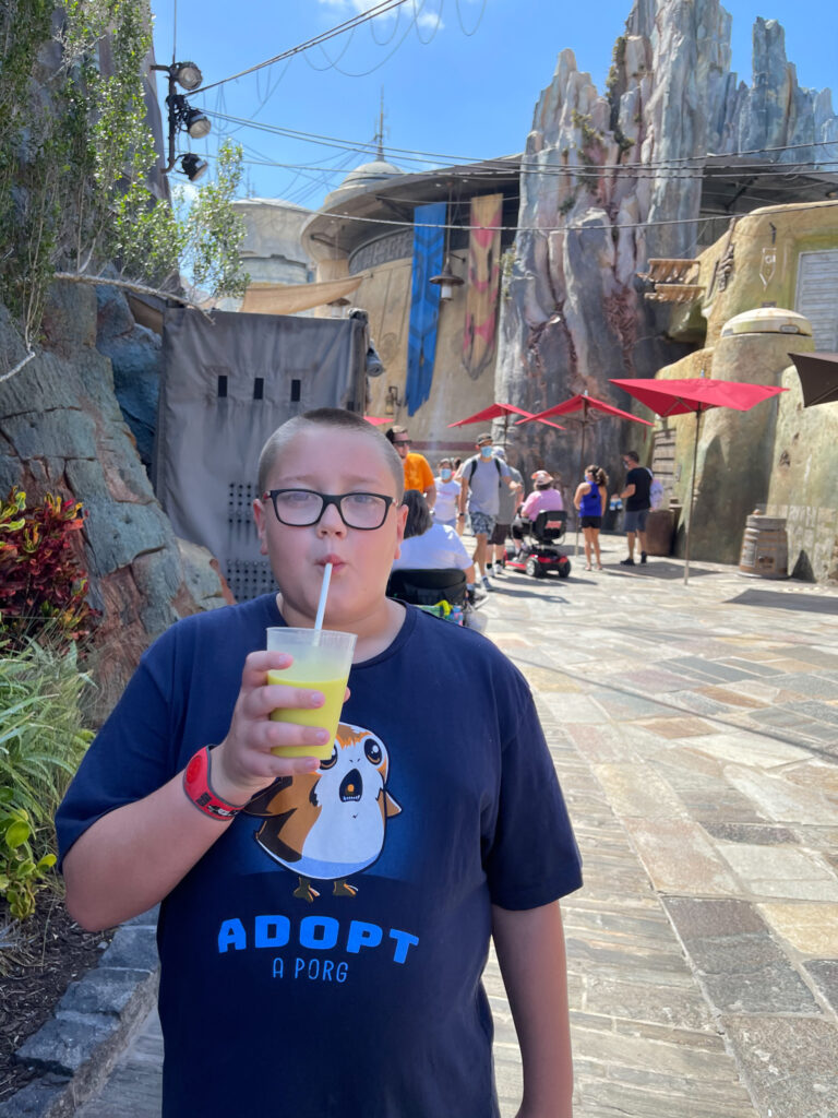  A boy wearing glasses and an ADOPT A PORG shirt drinks a green beverage with a straw in a sci-fi themed Disney World area—a fun spot for families, including those visiting Disney World with autistic kids. Rocky structures and visitors fill the background. 