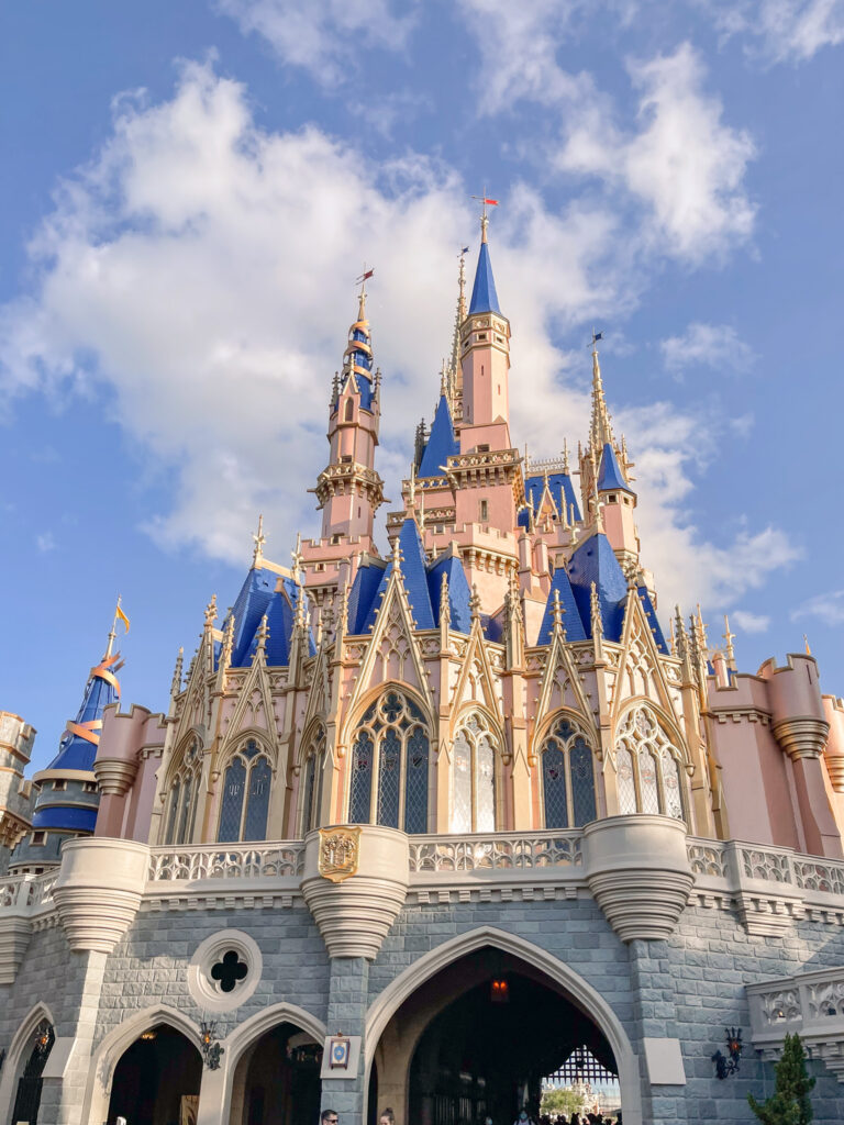  Cinderella Castle at Disney World, a magical sight for families, including those visiting Disney World with autistic kids, features blue and gold spires, pink walls, and intricate architectural details under a partly cloudy blue sky. 