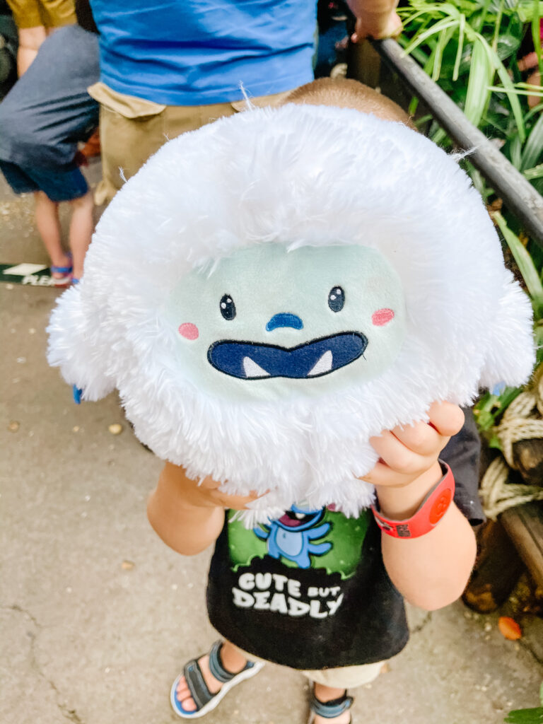  A child at Disney World with autistic kids holds a fluffy white yeti plush toy in front of their face, obscuring their head. The child wears a black shirt with a cartoon yeti and “Cute But Deadly,” sandals, and a red wristband. Others stand nearby. 